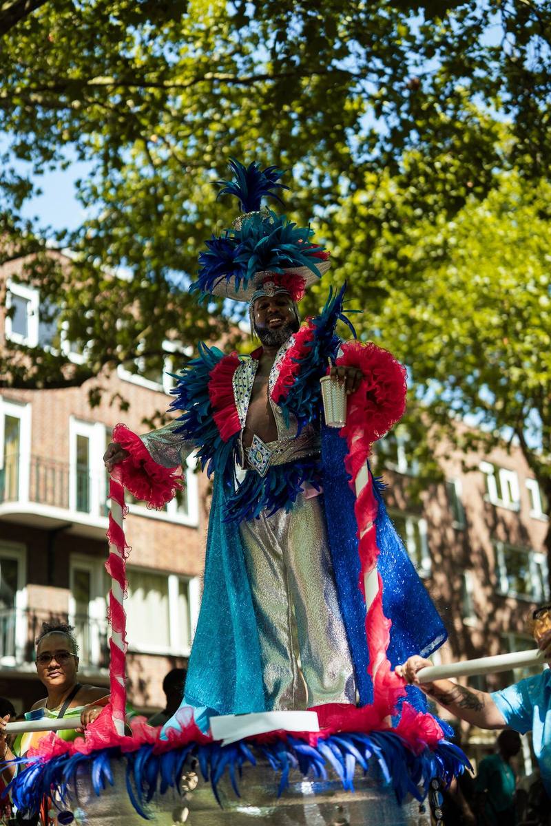 Een man in een blauw-rood kostuum met een hoge verentooi staat boven de stoet, met rood-wit gestreepte palen naast zich.
