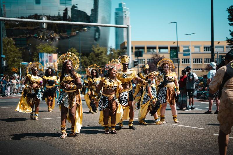 Een groep dansers in goud en luipaardprint trekt door een brede straat, met de Rotterdamse skyline erachter.